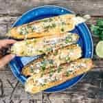 Four Elotes - grilled Mexican street corn - on a blue camping plate. A hand is reaching in to pick one up.