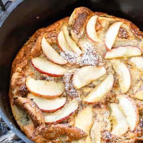 A baked apple pancake with sliced apples and powdered sugar sits in a black cast iron skillet over gray wood coals.