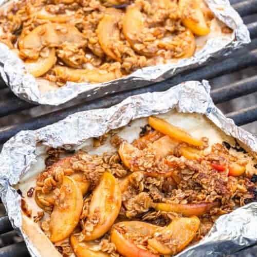 Two foil packets with baked sliced apples and a crumbly oat topping rest on a grill outdoors.