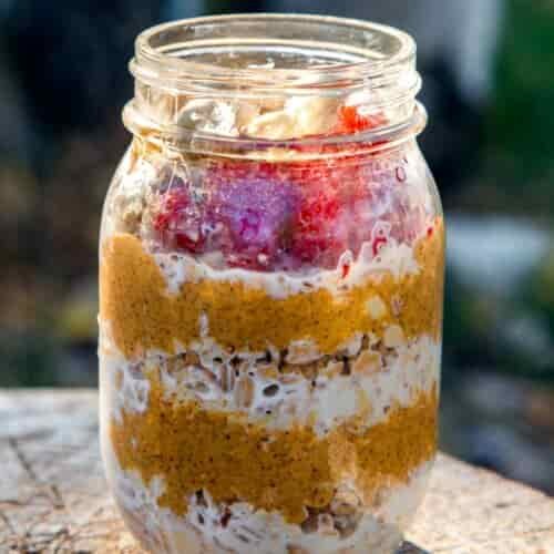 A mason jar with visible layers of oats, smooth nut butter, yogurt, and sliced strawberries rests on a wooden surface outdoors, with a blurred tree background.