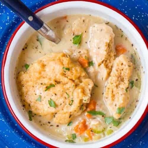 A white bowl with a blue edge and blue speckled plate holds creamy chicken and dumplings soup, featuring chunks of chicken, carrots, celery, herbs, and soft dumplings on a wooden table with a spoon resting inside.