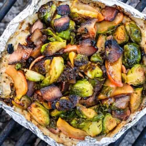 A foil packet on a grill grate contains Brussels sprouts, apple slices, and bacon pieces with charred bits and a roasted look.