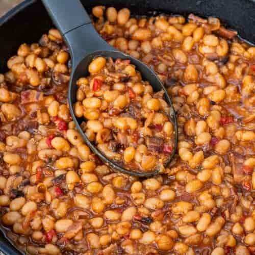 A black cast iron skillet filled with baked beans in a thick sauce sits on a rustic wooden surface, with a black serving spoon scooping up some of the beans.