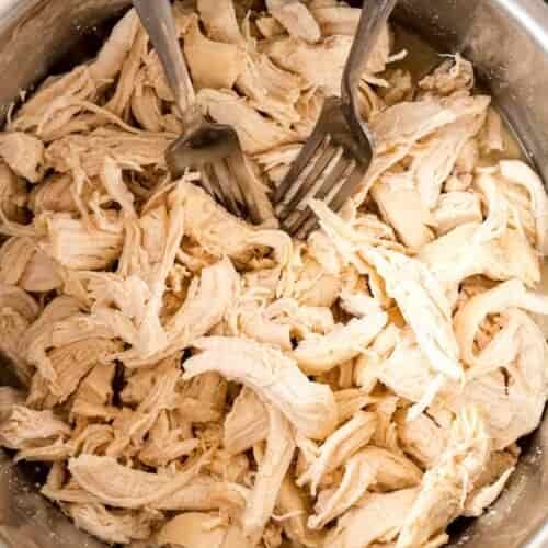 Two forks are shredding cooked chicken breast in a metal bowl.