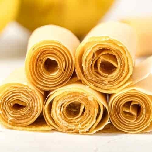 Five rolled sheets of light brown fruit leather are stacked in a pyramid shape on a white surface, with a blurred yellow object in the background.