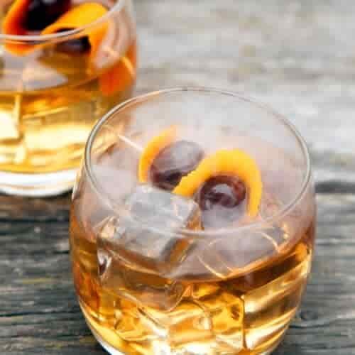 Two glasses of smoked old fashioned cocktails on the rocks with ice cubes, garnished with orange peel and two dark cherries each, sit on a weathered wooden surface with a hint of smoke visible above them.