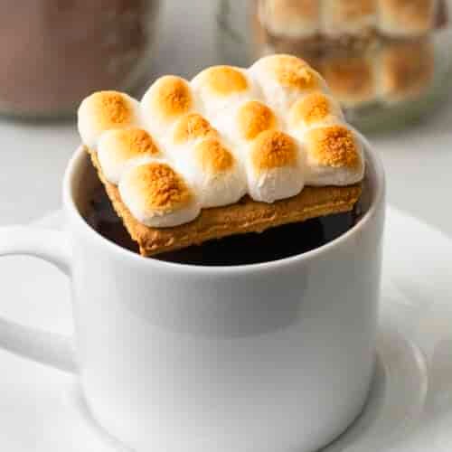 A white mug of hot chocolate is topped with a graham cracker and toasted mini marshmallows, with blurred jars in the background.