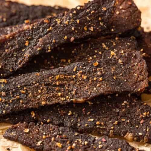 Close-up of several strips of dark, seasoned beef jerky on crumpled parchment paper, with visible spices and a textured, rustic appearance.