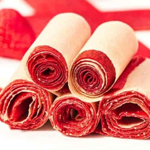 Five bright red, rolled strawberry fruit leathers with visible seeds are stacked on a white surface, each wrapped in light brown parchment paper, with blurred strawberries in the background.