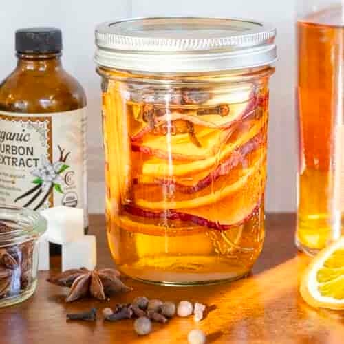 A mason jar with orange slices and spices on a wooden surface, surrounded by bottles, a bowl of star anise, sugar cubes, and an orange slice.